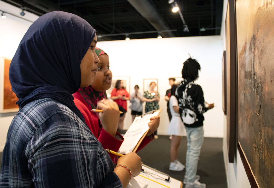 Two female students, one in a blue plaid shirt and the other in a red shirt, both wearing hijabs stand in an art gallery holding clipboards and pencils. They are looking at a painting on the wall, with other students and art pieces visible in the background.