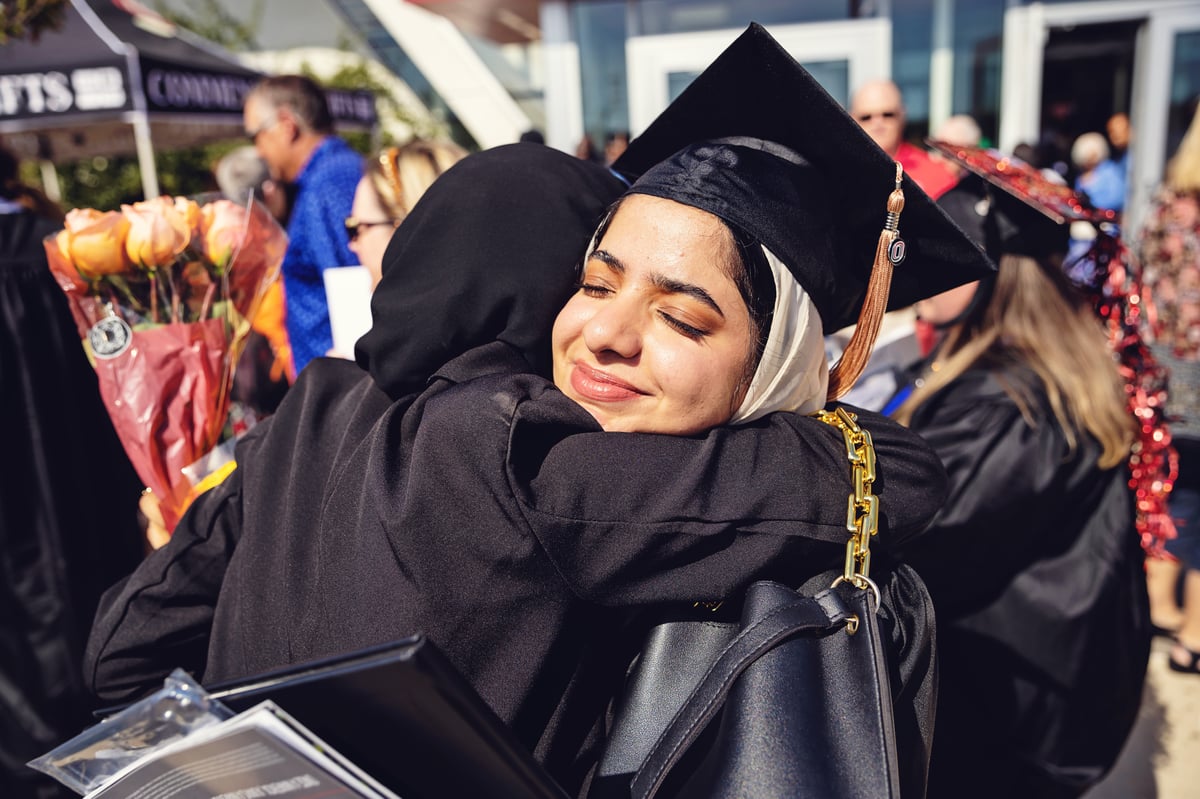 Girl smiling in her graduation cap and gown while hugging another person