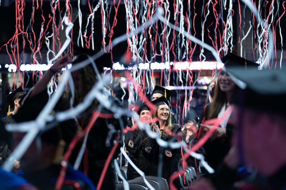 Graduates in caps and gowns smile and applaud as red, white, and black streamers fall from above during a commencement ceremony.
