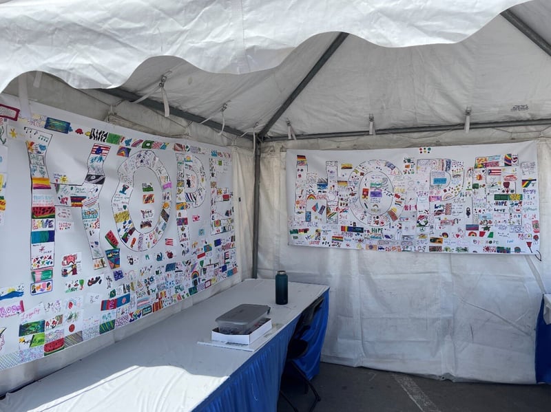 Image Alt text: SBMLC's booth at the 2024 Omaha Summer Arts Festival. Hanging from the tent frame are two large vinyl banners with the word 