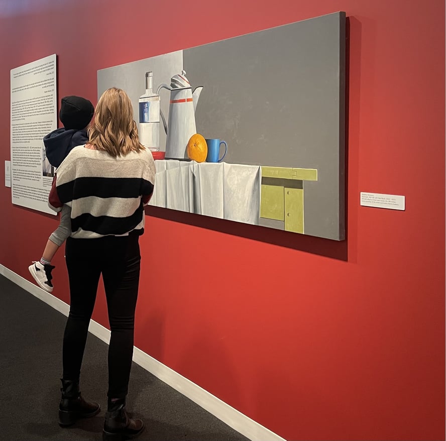 A woman is seen holding a child in front of a red wall. They both are looking at a large Stephan Namara painting.