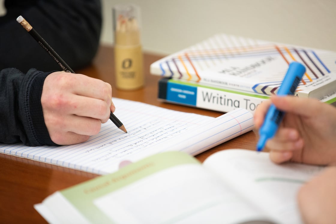 a close up of a table with text books, a hand in the background that is taking notes on a legal pad, and another hand in the foreground highlighting sections of a book