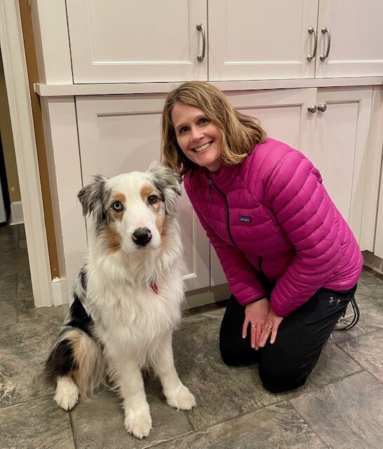 A woman in a pink jacket kneels on the floor next to a white long haired dog with black and tan spots. There are white cabinets behind them.