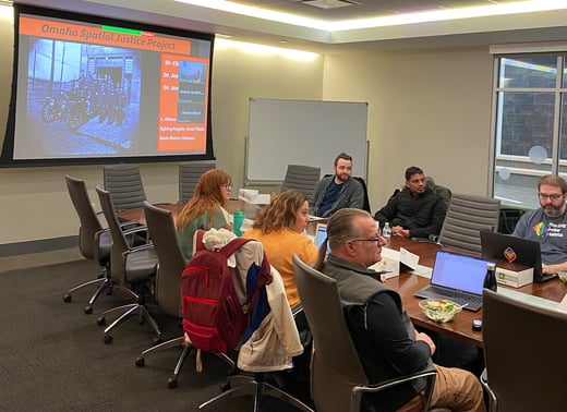 Students are sitting at a conference table, there is a PowerPoint presentation in the background