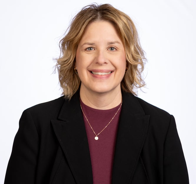 A smiling woman with medium length, wavy blonde hair, wearing a maroon round neck sweater with a black blazer and a necklace. 
