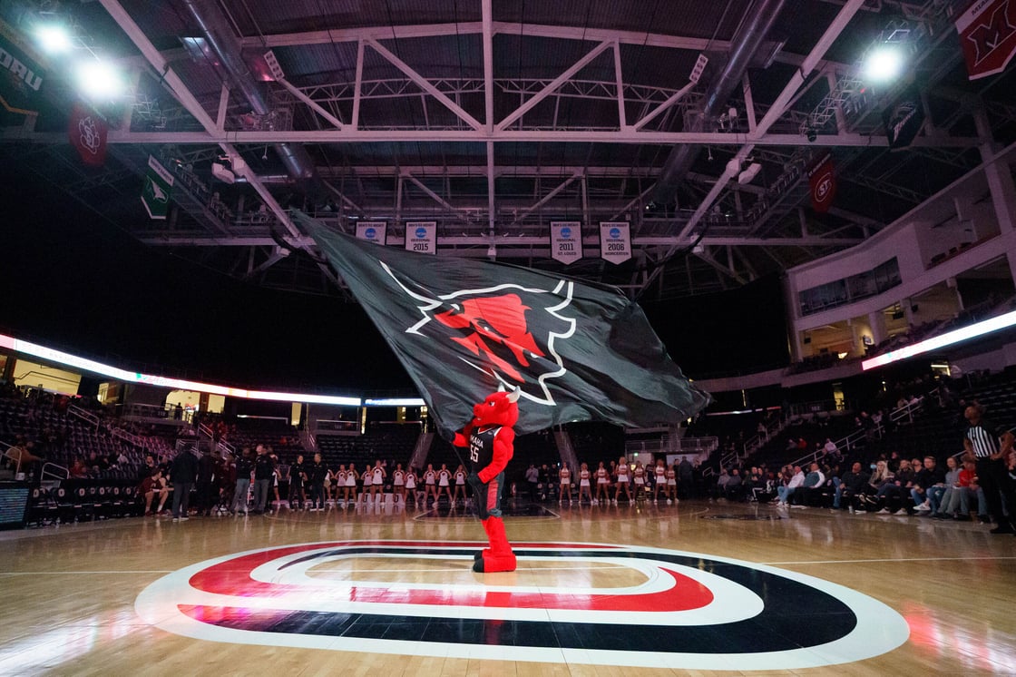 UNO's mascot, Durango, center court of Baxter Arena waiving UNO's athletic logo on a flag. UNO Dance and Cheer can be seen in the background.