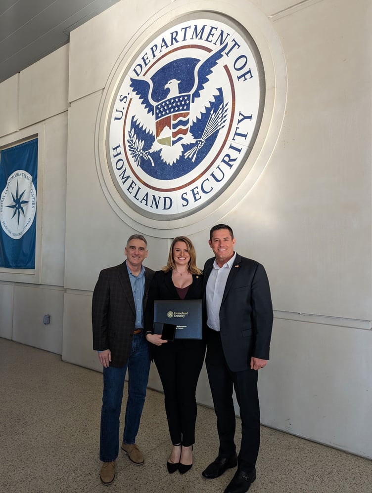 Col. John Persano, Emma, and SDSU's Lance Larson stand in front of a DHS seal.